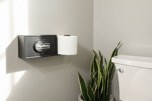 WipeWorks toilet paper holder and wet wipe dispenser mounted on a bathroom wall next to a toilet and plant.